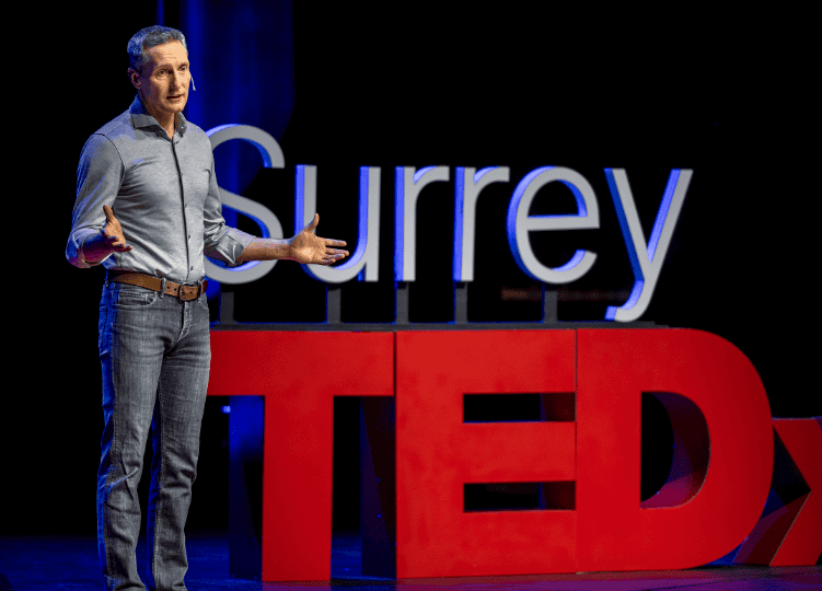 Michael Timms speaking on stage next to a large red TED sign, presenting his talk on claiming leadership power without relying on hierarchy.