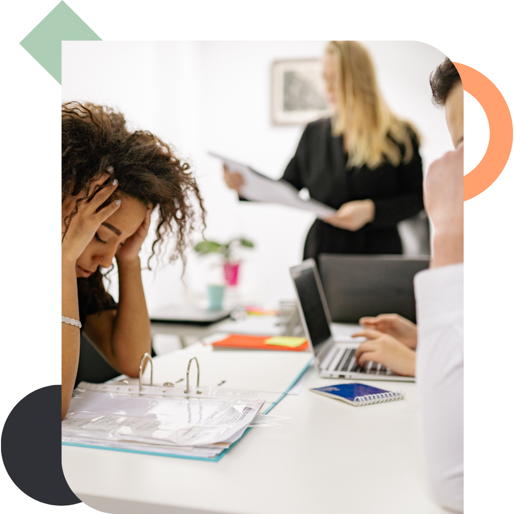 A woman with curly hair sits at an office table with her head in her hands, looking visibly stressed and overwhelmed, while colleagues work on laptops and review papers in the background, illustrating tension and hesitant workplace communication.