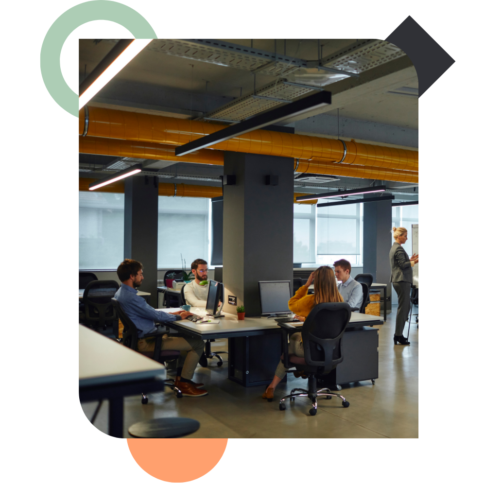 An open-plan industrial office with yellow overhead pipes, where a few employees work at their individual computer desks spread across a central pillar, capturing a disconnected corporate environment.
