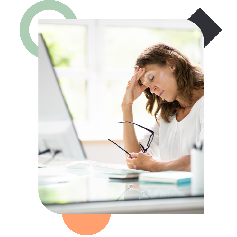 A woman sitting at her office desk with her eyes closed, holding her glasses in one hand and pressing her hand against her forehead in exhaustion, depicting burnout and severe workload capacity issues.