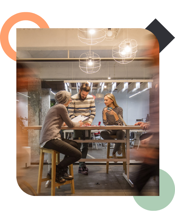 A group of three colleagues huddled around a high table in a brightly lit, modern office space discussing a document, while blurry figures of people walking past create a sense of constant motion and surface-level busyness.