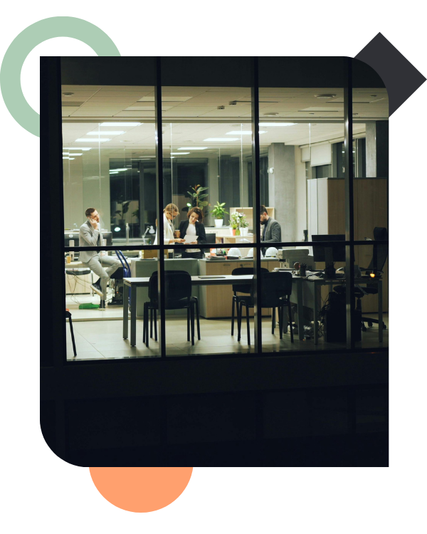 View through a large window of an office interior at night, showing a few employees still working late. One person sits on a desk talking on the phone while others huddle over paperwork, illustrating workplace pressure and extended hours.