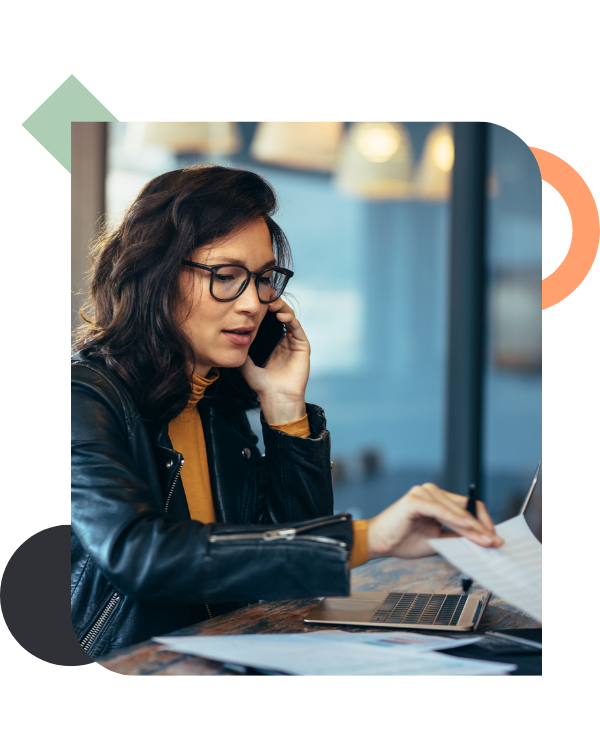 A woman with glasses and a black leather jacket sits at a wooden table, looking concerned while talking on a phone and flipping through paperwork with a laptop open in front of her, illustrating the heavy operational load placed on managers.
