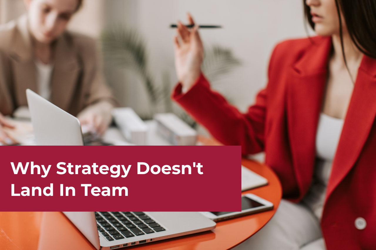 An article banner titled "Why Strategy Doesn't Land In Teams" features two professionals working at a bright orange round table. In the foreground, a woman wearing a vibrant red blazer holds a pen while gesturing during a conversation, with an open laptop and a smartphone sitting on the table in front of her. In the blurred background, another colleague in a tan blazer is focused on their work. A large, dark red rectangular overlay across the bottom left displays the text "Why Strategy Doesn't Land In Team".
