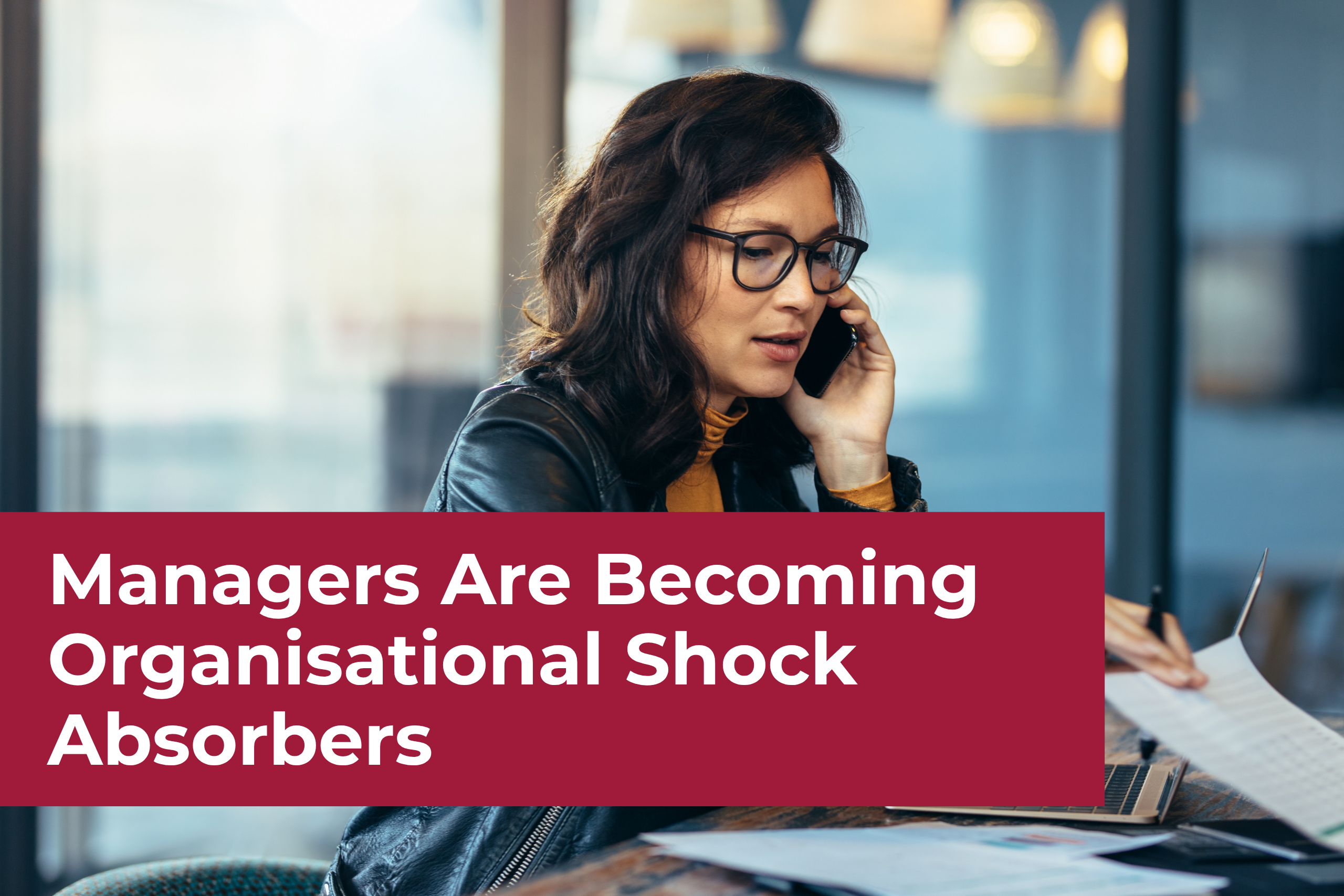 A focused female manager wearing glasses talking on her mobile phone while sitting at a desk with paperwork and a laptop. A prominent dark red banner displays the text, "Managers Are Becoming Organisational Shock Absorbers."